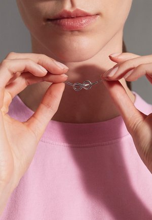 Silbernes Unendlichkeitshalsband, das in Händen gehalten wird, mit einem glatten Design und glänzender Oberfläche, vor einem sanften rosa Textilhintergrund.