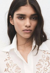 Blusa blanca con detalles intrincados de encaje en el lado izquierdo, con cuello clásico y una textura de tela suave y ligera.