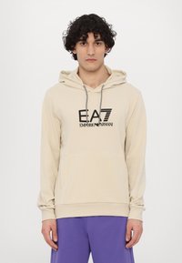 Felpa con cappuccio beige in tessuto morbido, dotata di tasca a marsupio, lacci a contrasto e logo nero stampato sul davanti.