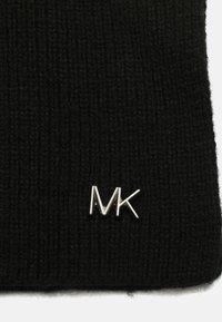 Musta neulepipo, jossa on kyynärpituinen rakenne ja hopeinen metallinen logo, jossa lukee "MK" etupuolella. Pehmeä ja joustava materiaali.