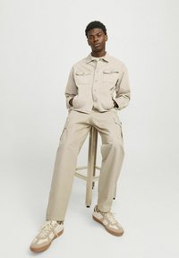 Beige denimjakke med to brystlommer og matchende cargo-bukser. Lyse sneakers med teksturerede detaljer, stående på en skammel.