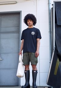 Camiseta de algodón negra con un gráfico circular blanco, pantalones cortos verdes tipo cargo, botas de goma negras y una bolsa de lona blanca. De pie junto a una puerta.