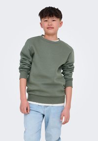 Sudadera verde de cuello redondo con puños y dobladillo acanalados, usada sobre una camisa blanca, combinada con jeans de color azul claro.
