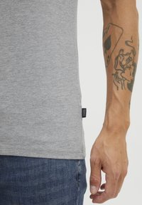 Tricou gri din bumbac, cu mâneci scurte, ajustat, având o mică etichetă neagră pe lateral. Purtați cu blugi din denim albastru.