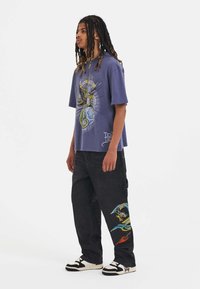 Lilla grafisk t-shirt med korte ærmer har et farverigt design. Sorte cargo-bukser med drage- og flammebroderi. Sorte og hvide sneakers.