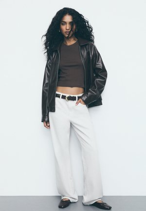 Giacca in pelle marrone con zip sopra un top corto marrone, abbinata a pantaloni larghi bianchi e scarpe basse scure. Design semplice e colori neutri.