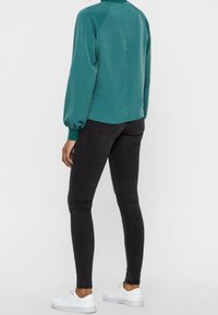 Blusa teal con maniche a sbuffo, dettaglio della cucitura posteriore, abbinata a jeans neri slim-fit e sneakers bianche. Tessuto morbido e forma aderente.
