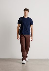 T-shirt navy a girocollo, pantaloni larghi marroni e sneakers bianche. Il modello porta una borsa a tracolla nera. Design semplice e vestibilità comoda.