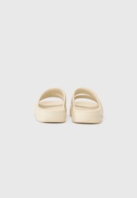 Beige slide sandals with an open back, two curved straps, and a flat sole. Smooth texture, simple design, suitable for casual wear.
