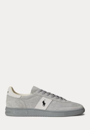 Basket basse en daim gris clair avec des lacets gris, un empiècement latéral blanc orné d’un logo noir de joueur de polo et une semelle en caoutchouc grise.