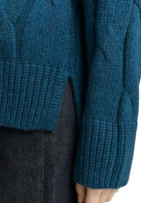 Pull en tricot teal avec un design en relief et des câbles, des poignets côtelés et des fentes sur le côté. Le matériau paraît épais et chaud, idéal pour les temps plus frais.