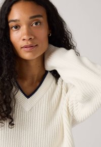 Cremefarbener V-Ausschnitt-Pullover mit gerippter Textur und dunkelblauem Akzent am Ausschnitt. Ein Model mit lockigem Haar posiert, mit der Hand am Hals.