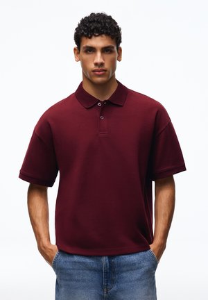 Mladý muž s kučeravými vlasmi, oblečený v bordové polo tričko s krátkym rukávom a modré džínsy, stojaci s rukami v vreckách na bielom pozadí.