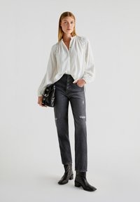 Blusa bianca a maniche lunghe con texture, con un'apertura a bottoni, abbinata a jeans neri sbiaditi a vita alta e stivaletti neri con tacco. Borsa nera.