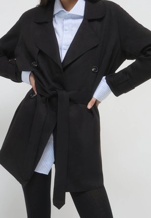Vrouw draagt een zwarte trenchcoat met riem over een lichtblauw gestreept overhemd met knopen en zwarte broek, handen in de zij.