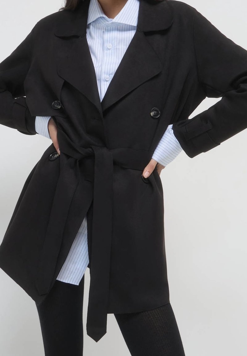Femme portant un trench-coat noir ceinturé par-dessus une chemise à rayures bleu clair boutonnée et un pantalon noir, mains posées sur les hanches.