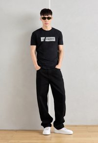 Black t-shirt with a white graphic, loose black pants, and white sneakers. Accessorized with green sunglasses. Wood floor and gray backdrop.