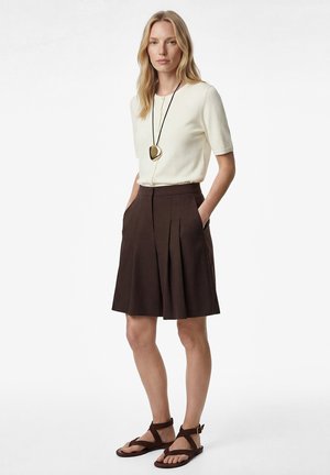 Vrouw draagt een crèmekleurige top met korte mouwen, een bruine geplooide rok tot op de knie, een zwarte hangerketting en bruine sandalen met bandjes, staand met handen in de zakken.