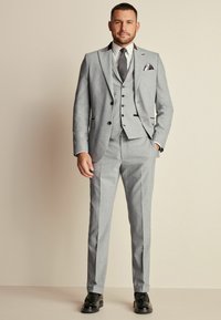 Traje gris de tres piezas con patrones sutiles, que incluye una chaqueta, un chaleco y pantalones ajustados, complementado con una camisa blanca y zapatos negros.