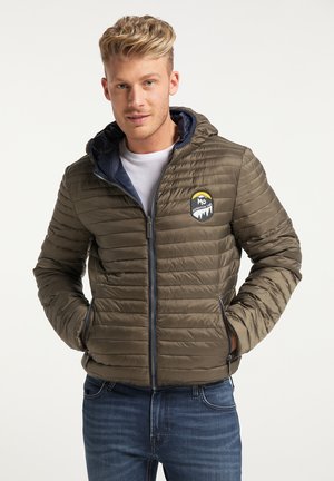 Jeune homme aux cheveux blonds courts portant une veste matelassée à capuche verte et un jean bleu, posant les mains dans les poches.