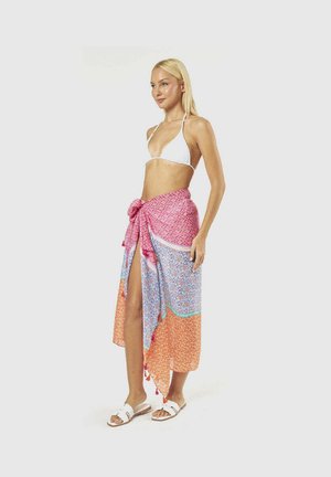 Donna con top bikini bianco, sarong colorato a fantasia annodato in vita e sandali bianchi in piedi su sfondo semplice.