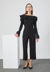 Svart off-the-shoulder topp med volangkant, lange ermer med utsvingte mansjetter, sammen med høyhalsede svarte bukser og svarte hæler.