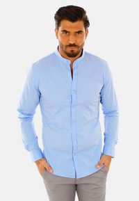 Camicia azzurro chiaro a maniche lunghe con colletto rialzato, texture sottile, chiusura frontale con bottoni e design aderente, abbinata a pantaloni grigio chiaro.