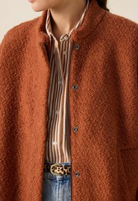 Veste texturée orange rouille avec un col montant, boutons-pression, et une chemise rayée contrastante en dessous, accompagnée d'une ceinture imprimé léopard.