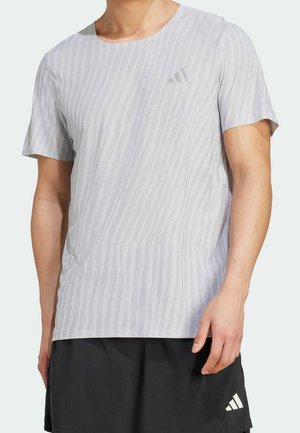 Mann trägt ein hellgraues, strukturiertes kurzärmliges Sportshirt und schwarze Shorts mit weißem Logo auf dem linken Bein.