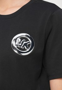 Fekete pamut póló, melynek mellkasán fényes ezüstszínű kerek embléma található. Rövid ujjú, kerek nyakkivágású, sima tapintású anyagból.