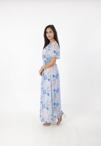 Robe maxi florale bleu clair avec manches courtes, dotée d'une taille cintrée et d'un tissu fluide. Comprend des motifs de fleurs roses et bleues. Talons transparents.