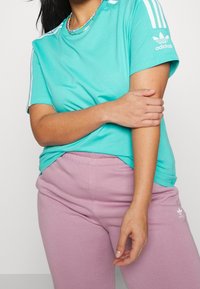 Persona vistiendo una camiseta turquesa de Adidas y pantalones deportivos morado claro de Adidas, tocándose el brazo izquierdo con la mano derecha.