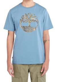 Muž v světle modrém tričku s logem maskáčového stromu a béžových cargo kalhotách, který stojí s uvolněnýma rukama podél těla.