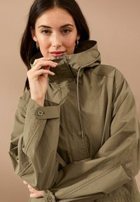 Olivgrüne Regenjacke mit hohem Kragen, verstellbarem Kapuzen und Druckknopfverschlüssen; verfügt über weite Ärmel und strukturierten Stoff.