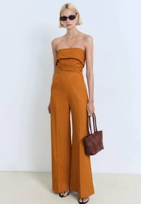 Strapless amber jumpsuit met rimpelingen bij de buste, wijde benen, gecombineerd met een kleine geweven bruine tas en donkere zonnebril. Gladde stof textuur.