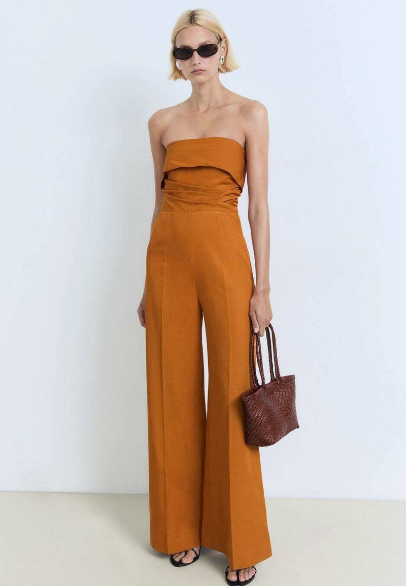 Strapless amber jumpsuit met rimpelingen bij de buste, wijde benen, gecombineerd met een kleine geweven bruine tas en donkere zonnebril. Gladde stof textuur.