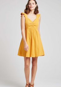 Robe en coton de couleur jaune moutarde avec un corsage ajusté, un col en V et des bretelles à volants. La jupe est évasée, arrive au genou et est en tissu texturé.