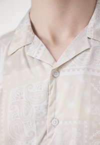 Kortærmet, beige skjorte med knapper, der har et paisley-mønster, glat stof, krave og to hvide knapper nær halsen.