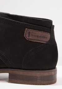 Bottine en cuir noir avec un patch marron étiqueté "bugatti", forme élégante, surface texturée et semelle en bois contrastante.
