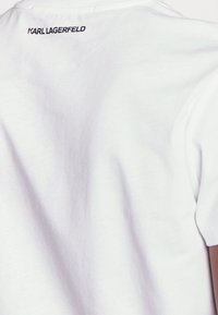 Vit bomullströja med rundad halsringning, korta ärmar och en svart "KARL LAGERFELD"-logotyp tryckt på den övre ryggen.