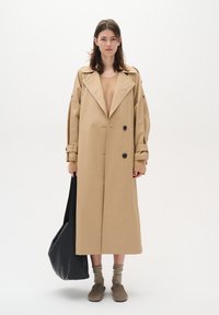 Beige trench coat with large lapels, double-breasted buttons, and relaxed fit. Black tote bag, brown slip-on shoes, and brown socks.