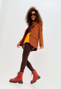 Rode suède enkellaarsjes met zwarte veters, rubberen zool, gecombineerd met een oversized bruine blazer, een oranje trui en een paarse panty.