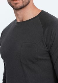 Dunkelgraues Langarmshirt mit strukturiertem Körper und glatten Ärmeln, ausgestattet mit einer kleinen Brusttasche und geripptem Kragen.