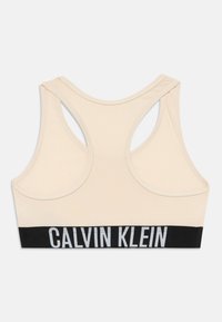 Bež sportski grudnjak s dizajnom na leđima. Ima crnu elastičnu traku s natpisom "CALVIN KLEIN" u bijelom na dnu. Glatka tekstura.