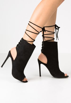 Czarne zamszowe botki na wysokim obcasie z odkrytymi palcami i bokami, ozdobione krzyżującym się sznurowaniem i nowoczesną, smukłą sylwetką.