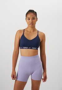 Mørkeblå sports-bh med V-hals og tynne stropper, med et hvitt Nike-logo bånd. Kombinert med høy midje lilla shorts.