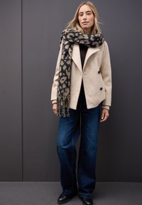 Beige dobbeltbreasted frakke, sort top, brede blå jeans og et mønstret sort og beige tørklæde med frynser. Sorte ankelstøvler.