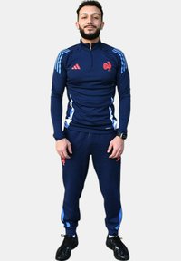 Chándal deportivo azul marino con un top de media cremallera, detalles rojos y franjas laterales azules; combinado con pantalones a juego y zapatillas deportivas negras.