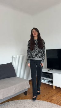 Grå genser med leopardmønster, svarte bukser og svarte slip-on sko. Står i en stue med sofa, radiator og TV-benk.