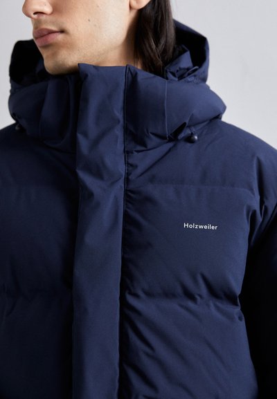 Doudoune bleu marine avec un col haut, fermeture éclair à l'avant et un logo discret sur la poitrine. Elle présente une texture lisse et mate ainsi que des bords arrondis.